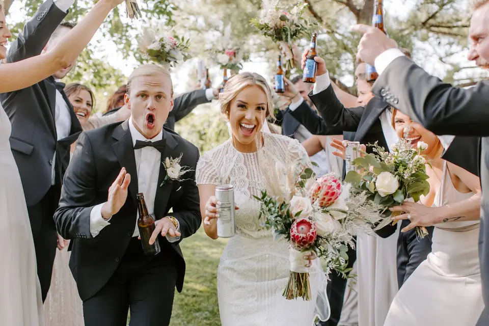 Bride and groom smiling as guests toss flowers and raise beer bottles around them