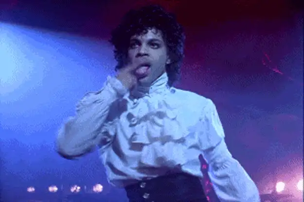 Animated GIF of a performer with curly hair in a ruffled white shirt on a purple-lit stage