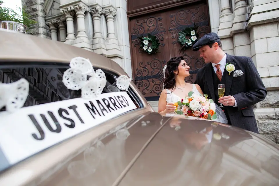Bride and groom beside a Just Married sign on a car outside a church, holding a champagne glass and bouquet