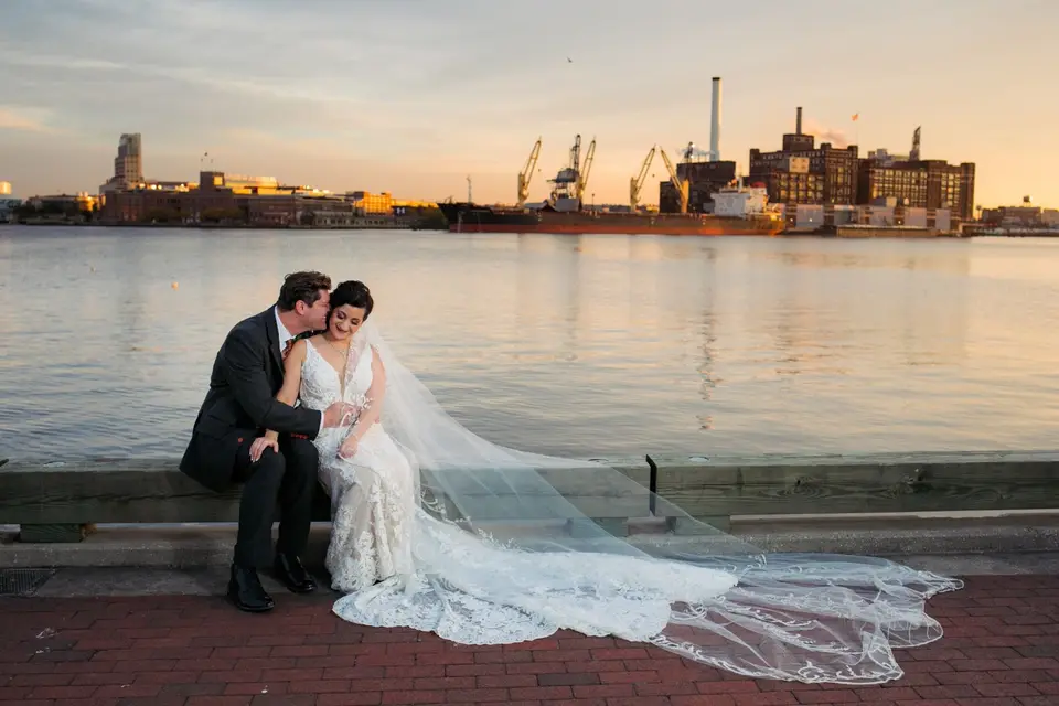 Bride and groom sitting on a waterfront seawall at sunset with her long lace veil spread behind them