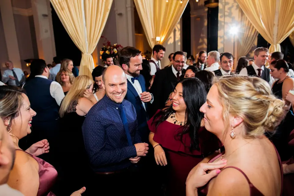 Smiling wedding guests dancing close together on a ballroom dance floor under draped curtains