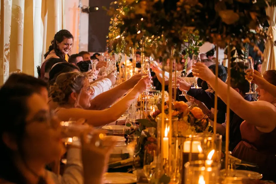 Wedding guests raising glasses in a toast across a long candlelit table with tall centerpieces