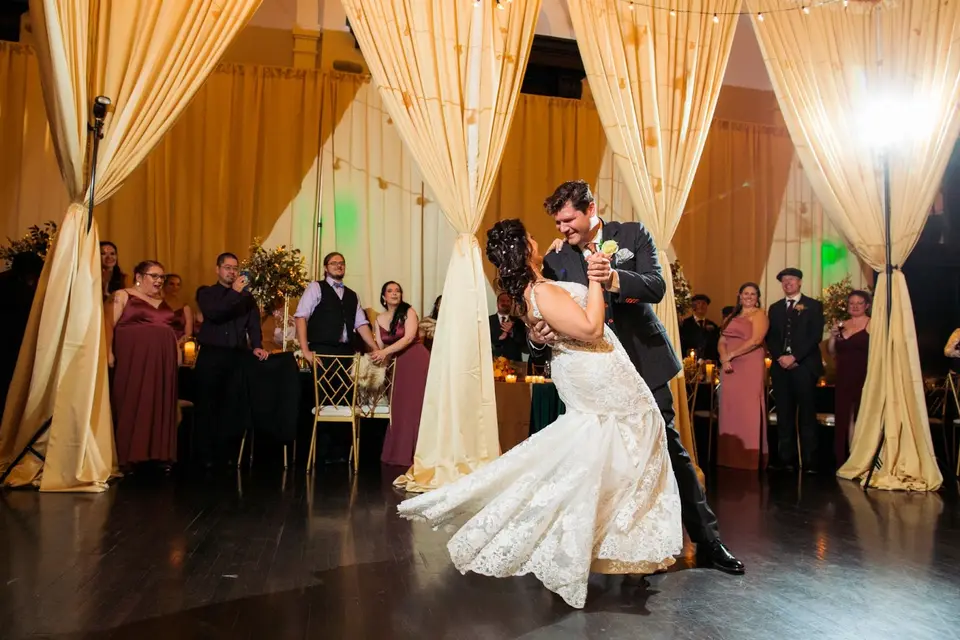 Bride and groom sharing a first dance in a ballroom with draped curtains while guests look on