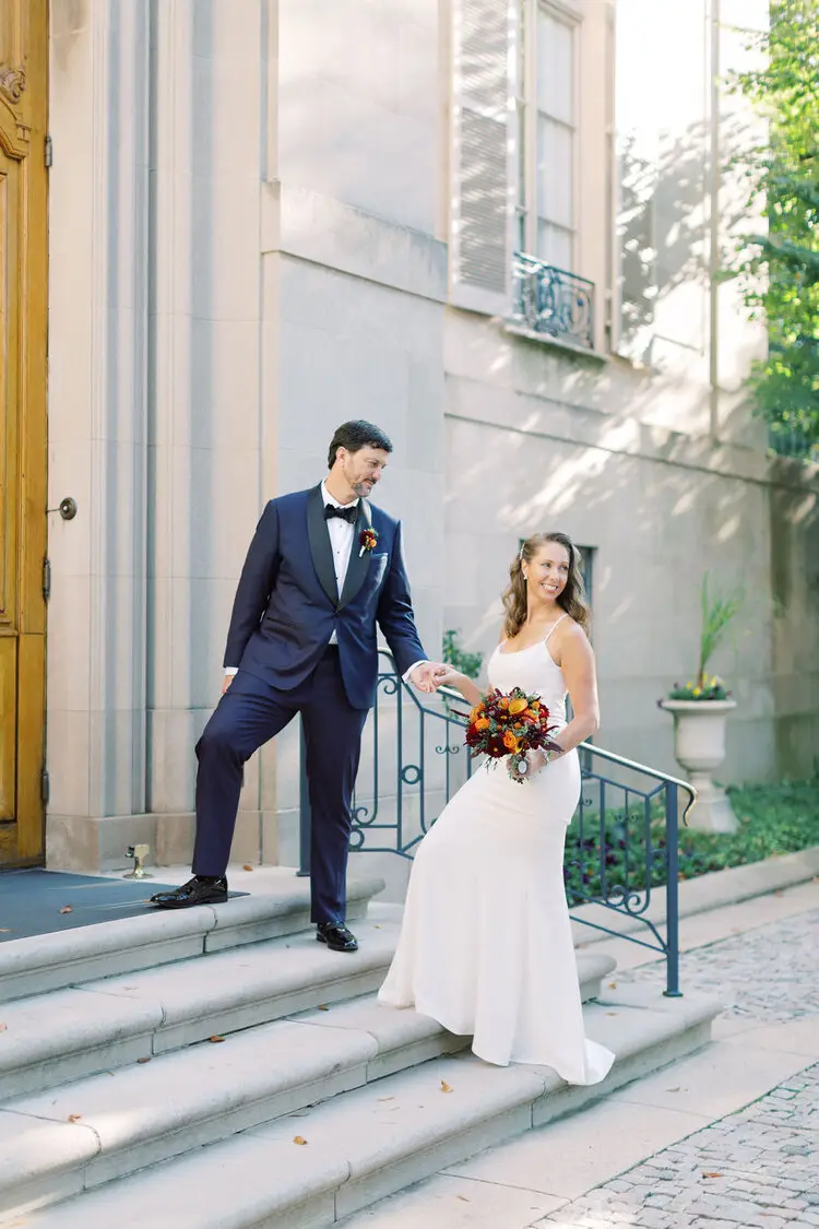 Groom in a navy suit leading the bride in a long white dress down stone steps, she holds an autumn bouquet