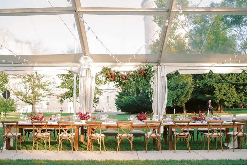 Long wedding reception table with wooden chairs and floral centerpieces under a clear tent on a lawn