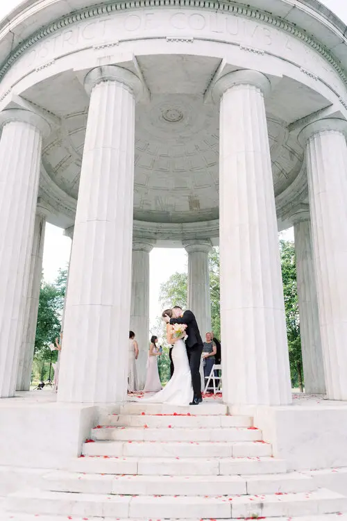 Couple embracing at the top of the steps inside a white columned memorial rotunda, bridesmaids standing nearby