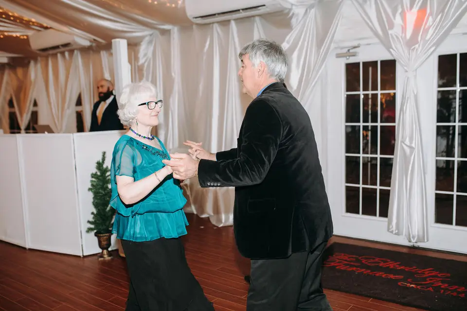 Older man in a velvet jacket dancing with a woman in a teal top, DJ booth visible behind them