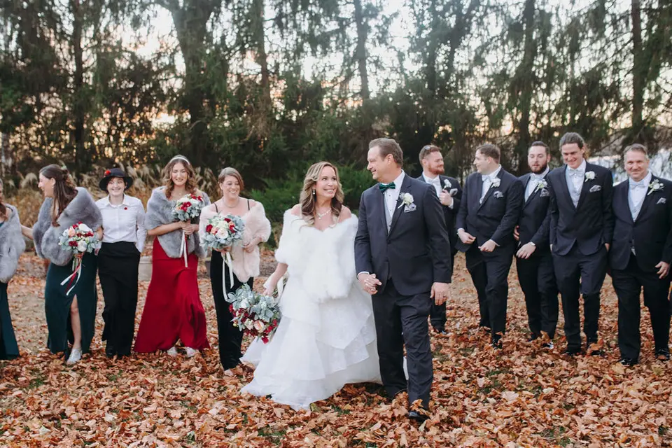 Bride in white fur wrap and groom walking hand in hand with bridesmaids and groomsmen over fallen autumn leaves