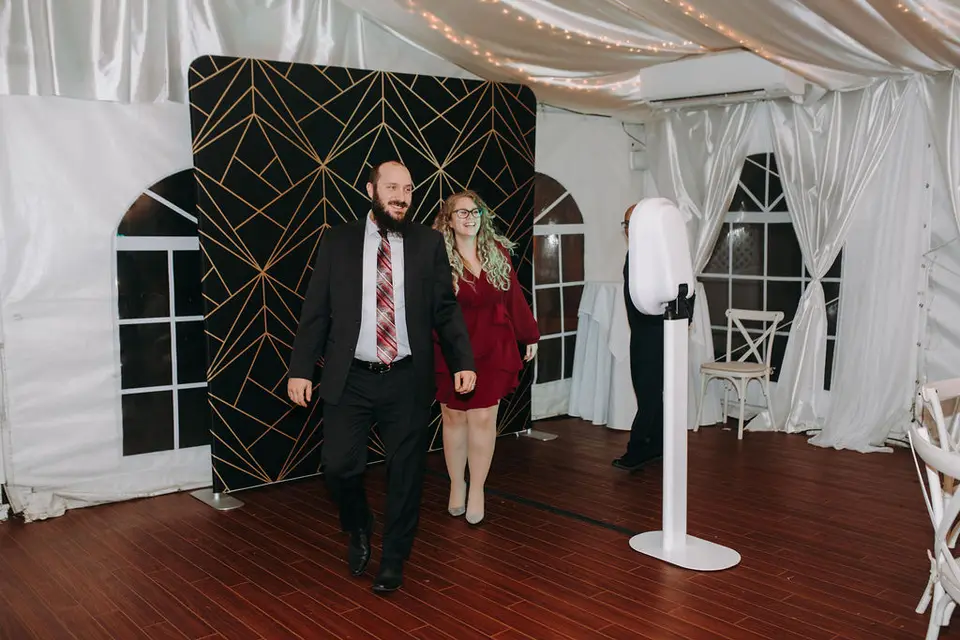 Man in a red striped tie and woman in a red dress walking past a photo booth and geometric backdrop