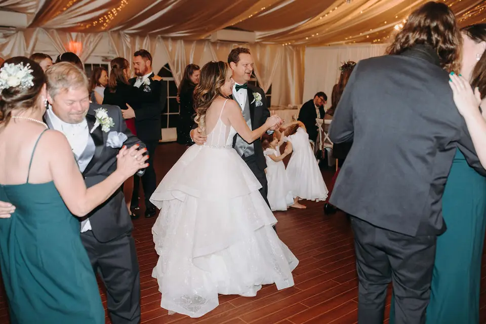 Bride and groom dancing with guests on a wood dance floor under a draped tent ceiling as flower girls watch