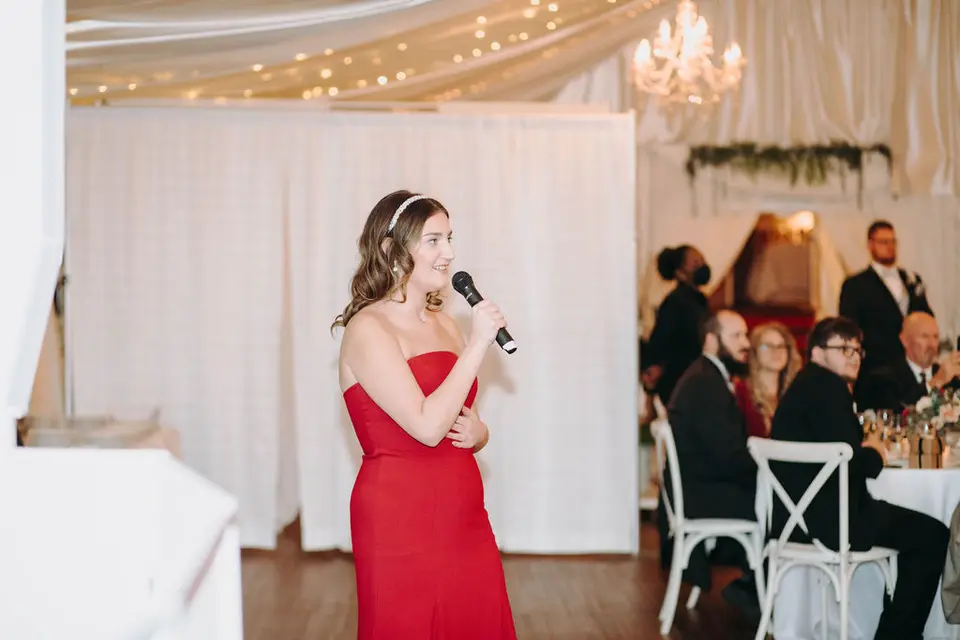 Bridesmaid in a red strapless gown giving a speech into a microphone in a tented reception with string lights