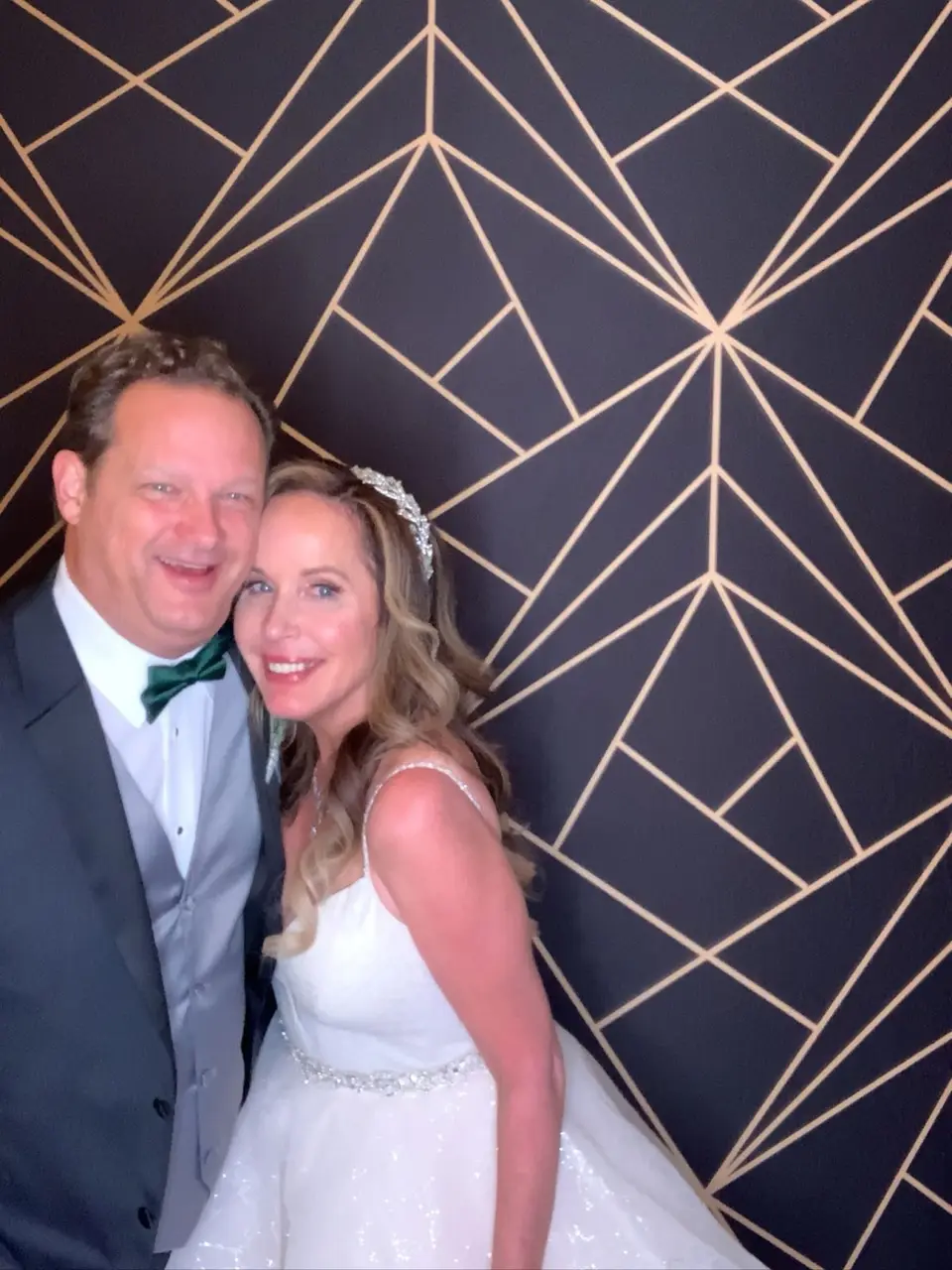 Groom in a green bow tie and bride in a white gown posing together in front of a geometric photo backdrop
