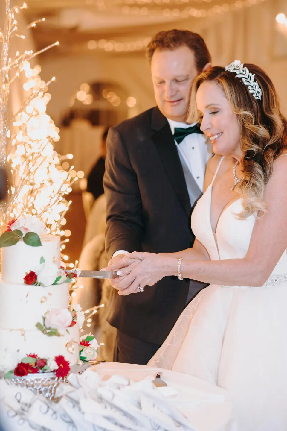 Bride and groom cutting a tiered wedding cake decorated with red roses beside a lit branch centerpiece