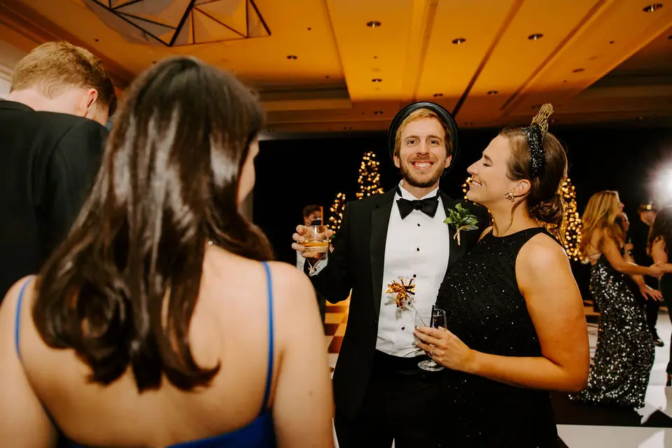 Smiling man in a tuxedo and woman in a black sequin dress holding drinks at a New Year's Eve wedding