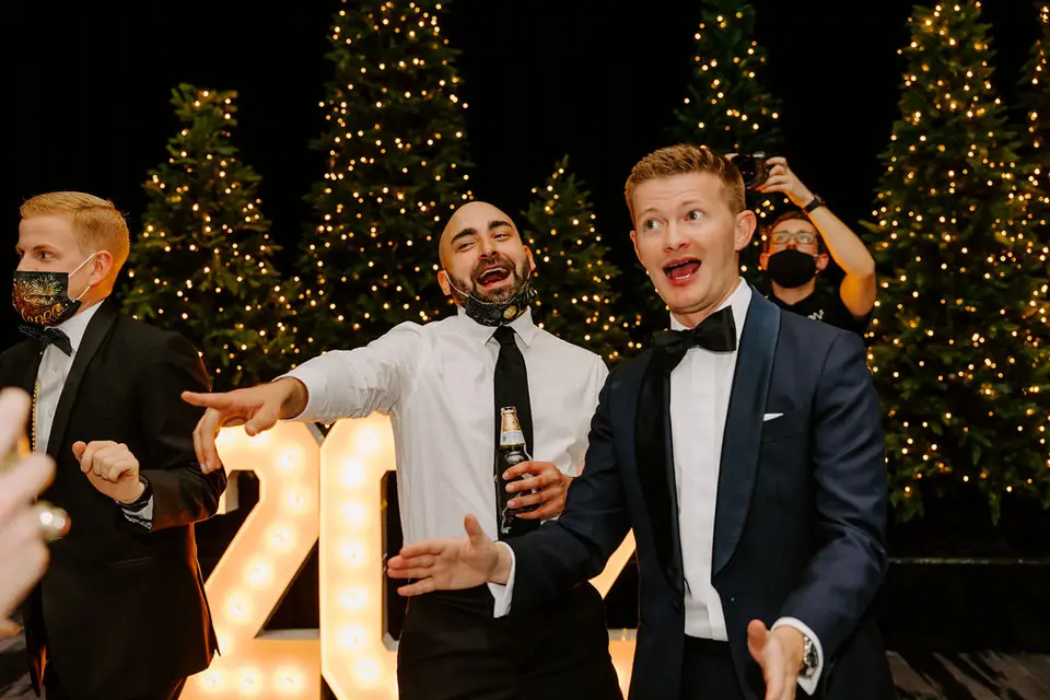 Two men in formal wear cheering and gesturing in front of lit Christmas trees and marquee numbers