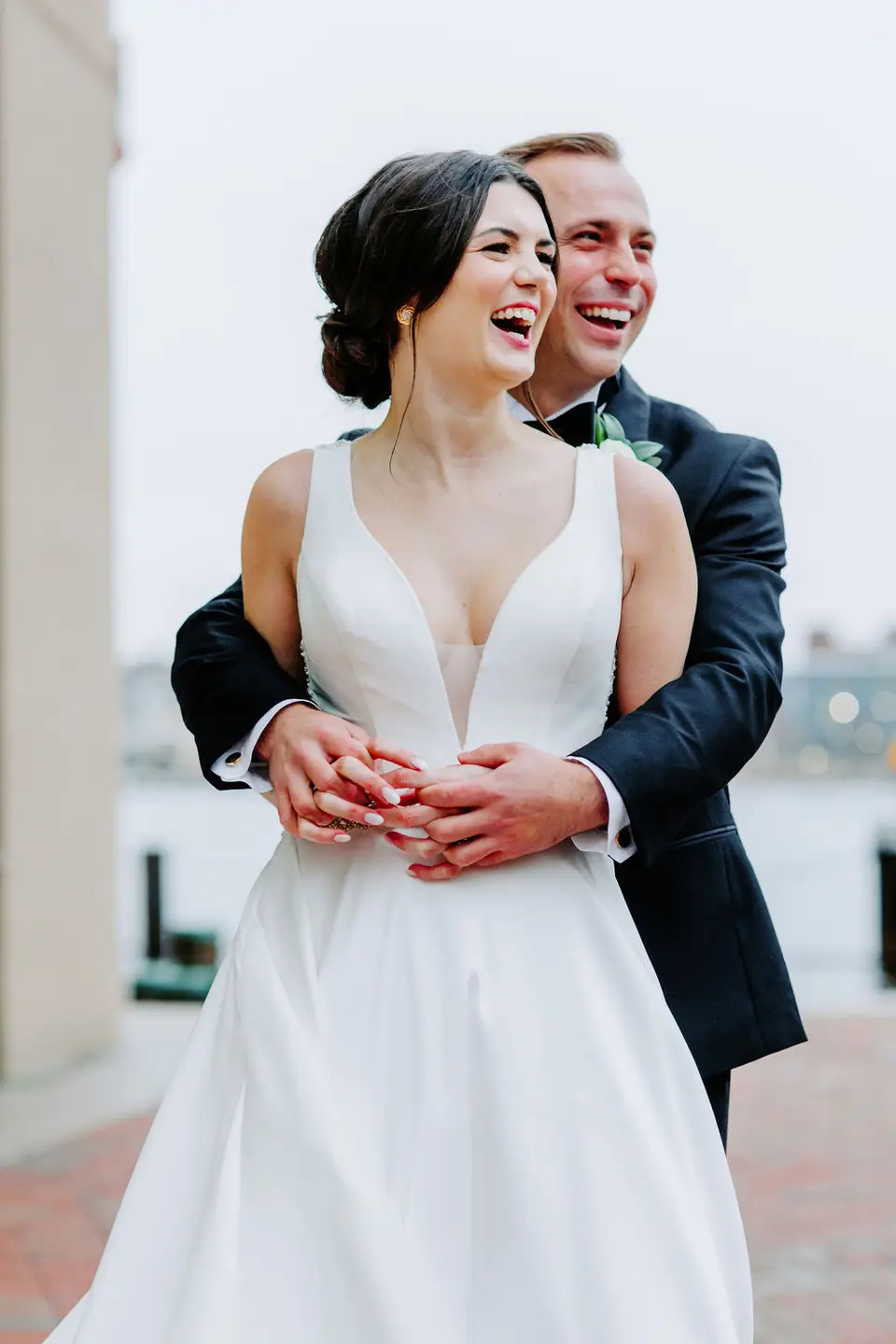 Bride and groom laughing together in an embrace on a brick waterfront walkway