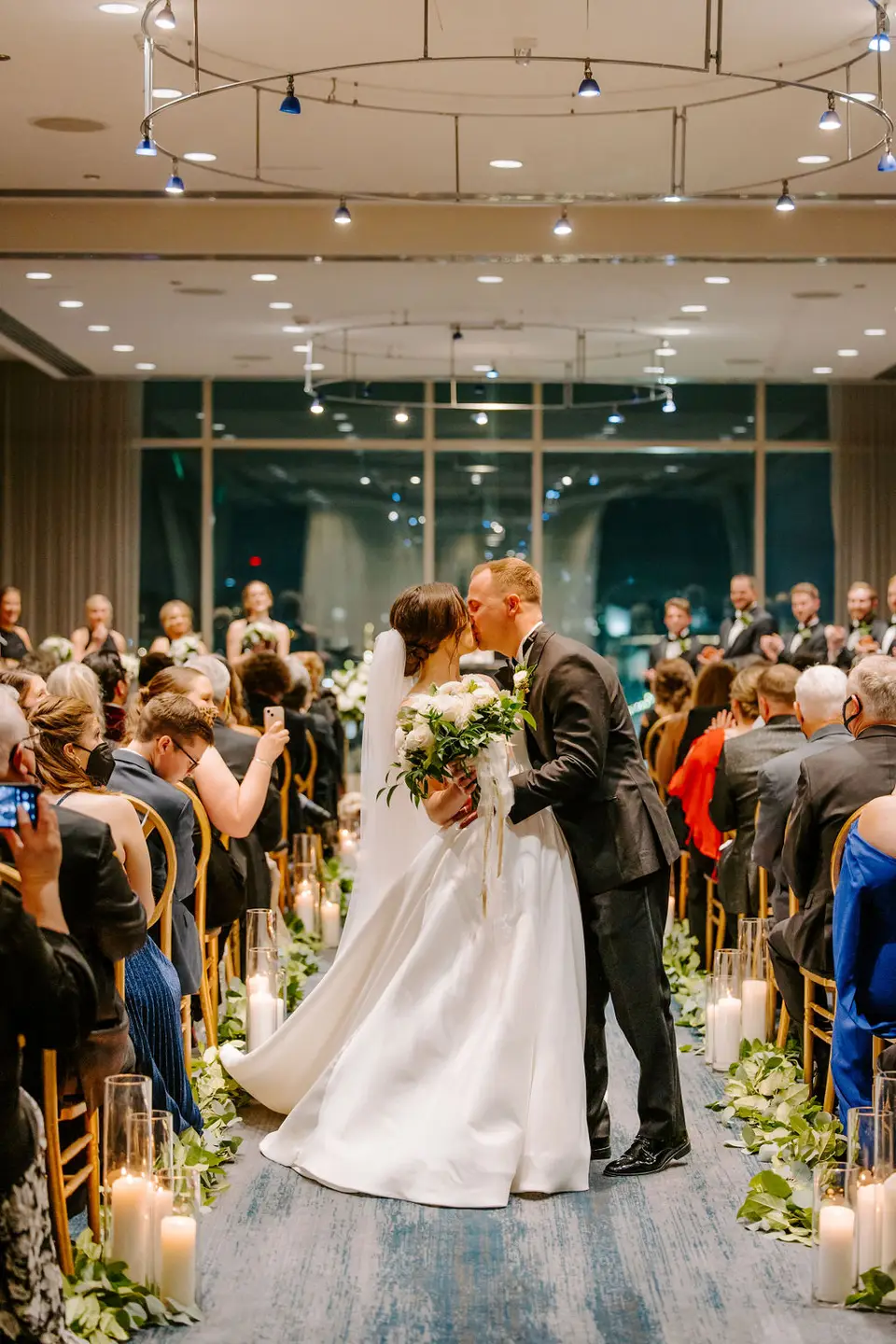 Bride and groom kissing in the aisle lined with candles and greenery while guests look on