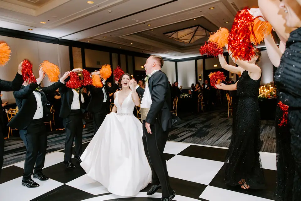 Bride and groom dancing on a black and white checkered floor as guests wave red and orange pom poms