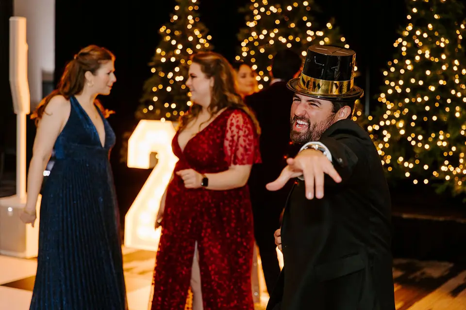 Bearded man in a gold top hat gesturing on the dance floor beside women in dresses and lit Christmas trees