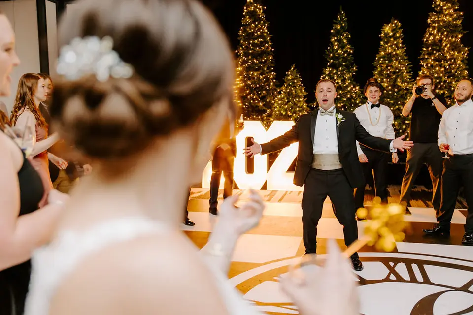 Groom in a tuxedo with arms out on a dance floor in front of lit Christmas trees and large marquee letters