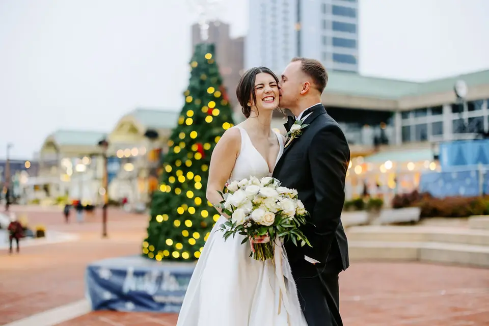 Bride and groom laughing as he kisses her cheek beside a lit Christmas tree at the Baltimore Inner Harbor