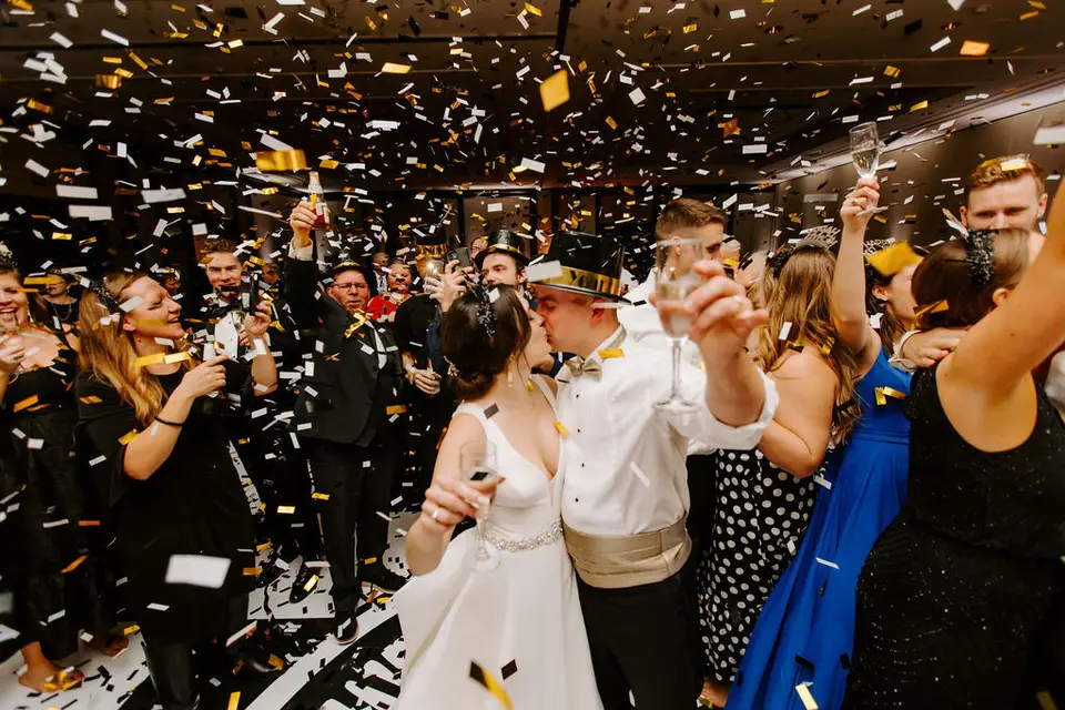 Bride and groom kissing surrounded by guests as gold and white confetti falls at a New Year's Eve reception