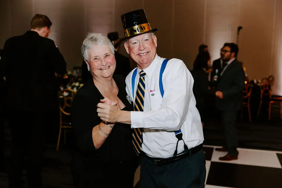 Older man in a gold New Year's top hat and suspenders dancing with a smiling older woman