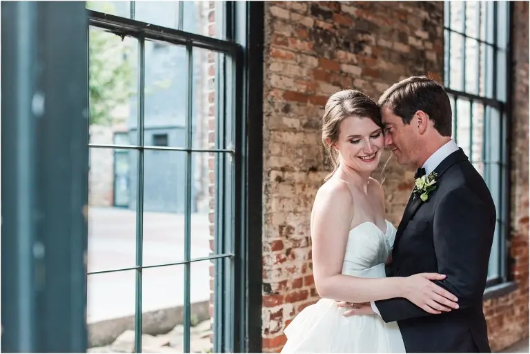 Bride and groom embracing by tall industrial windows and an exposed brick wall