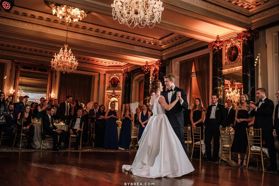 Bride and groom sharing a first dance in a ballroom with chandeliers as guests watch