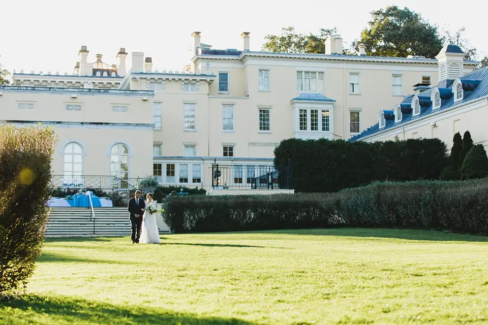Bride and groom walking on a lawn toward a large cream-colored mansion