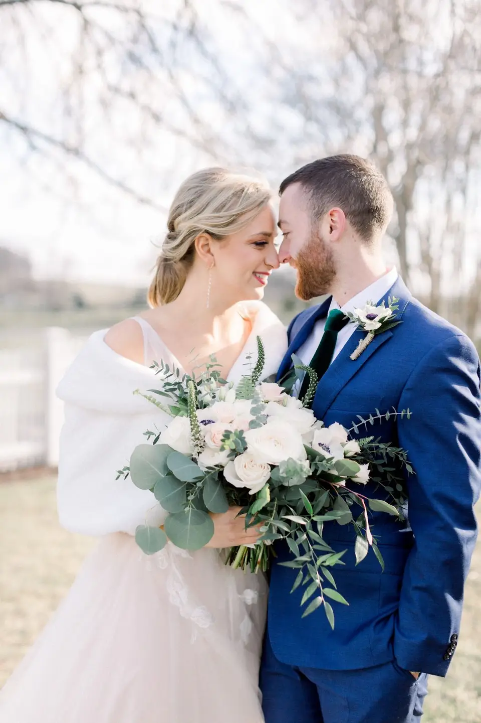 Bride and groom nuzzling close, holding a white and greenery bouquet outdoors