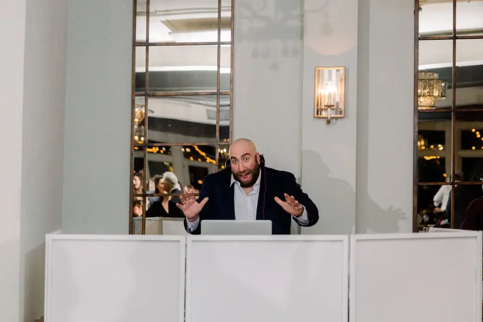 DJ in a dark blazer gesturing playfully behind a white DJ booth in a mirrored ballroom