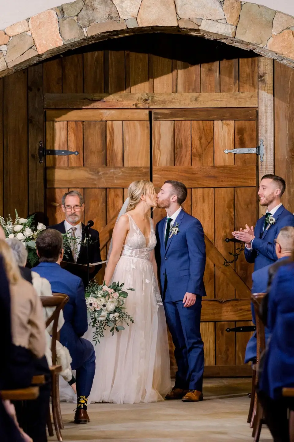 Groom kisses bride at the altar in front of a stone arch and wooden barn doors as the guests applaud