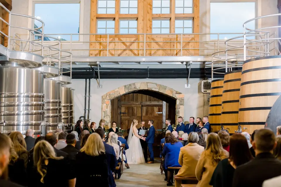 Couple exchanging vows at a winery ceremony between stacked wine barrels and steel tanks with guests seated
