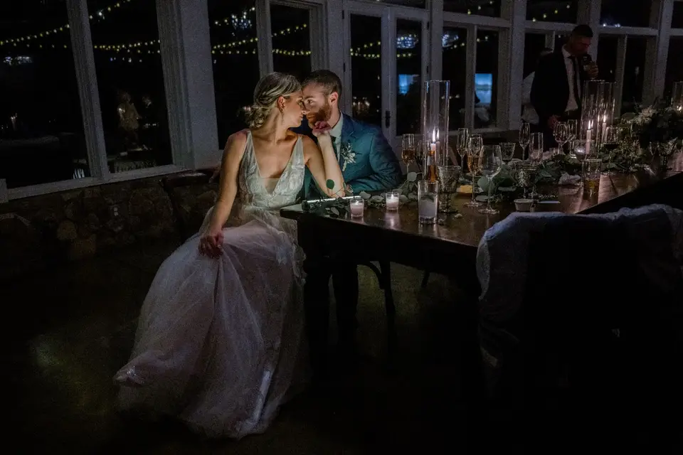 Bride and groom sitting close together at their candlelit head table in a dark room with window lights