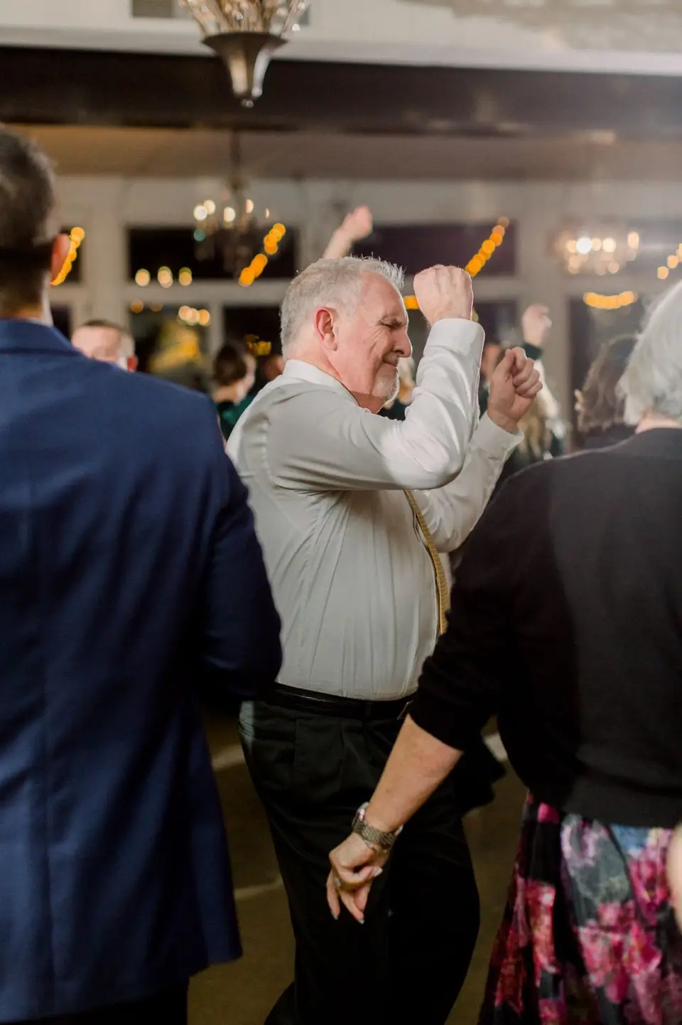 Older man in a gray shirt dancing with a fist raised among reception guests