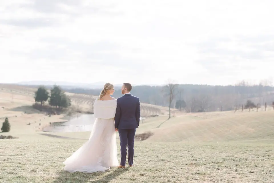 Bride in a white wrap and groom in a navy suit seen from behind, overlooking a vineyard and pond