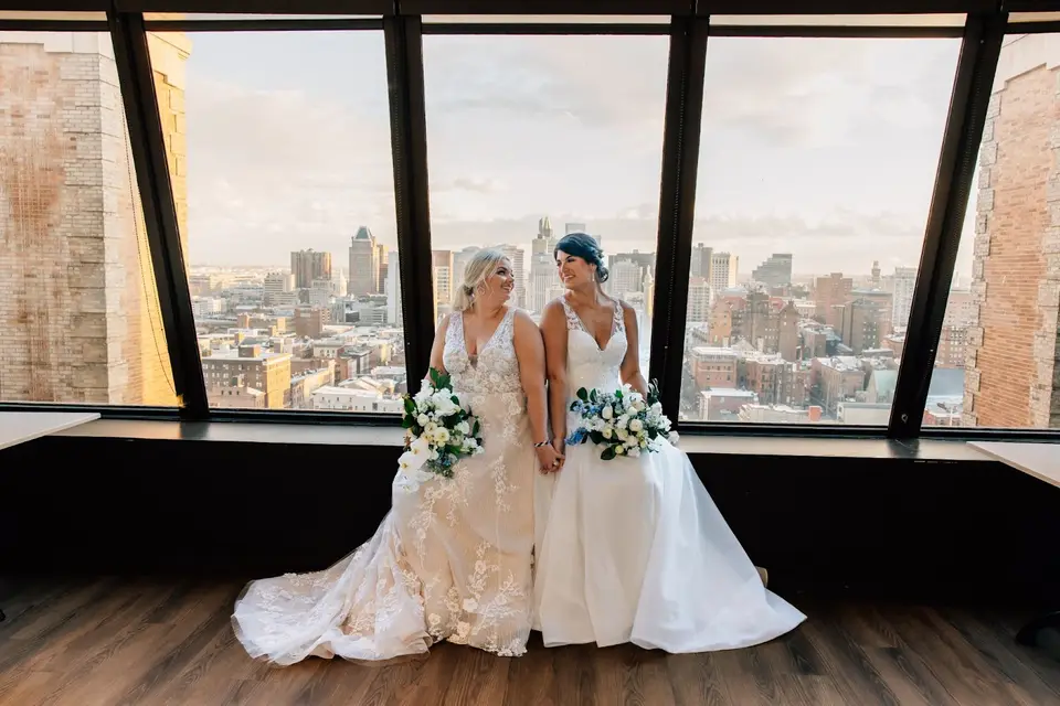 Two brides in wedding gowns sitting side by side by tall windows overlooking a city skyline
