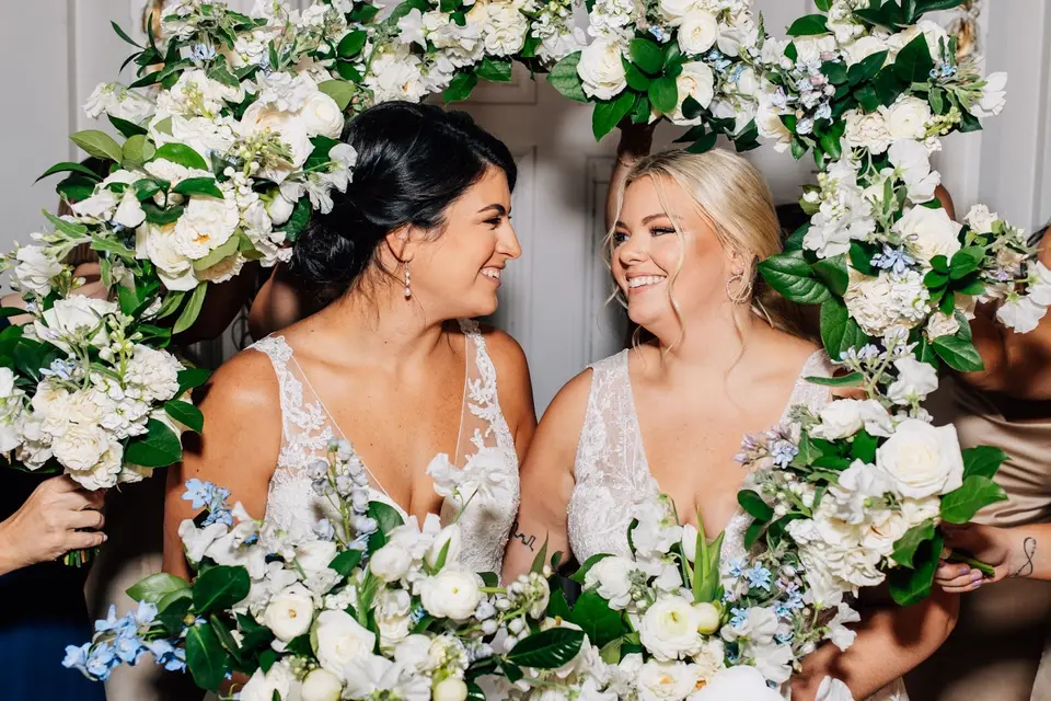 Two brides smiling at each other while surrounded by white and blue floral bouquets