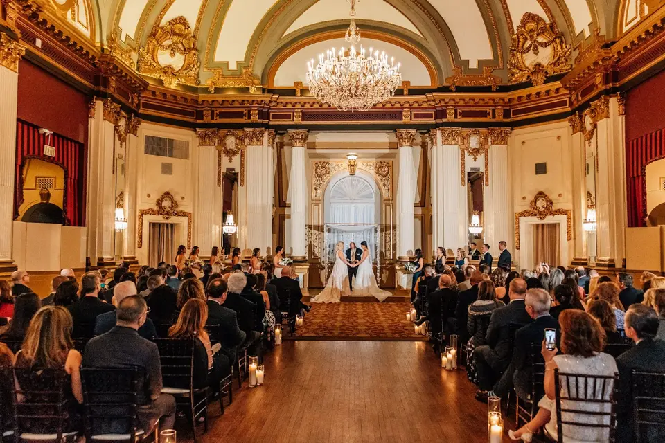 Wedding ceremony in a grand ballroom with a chandelier as guests watch two brides at the altar
