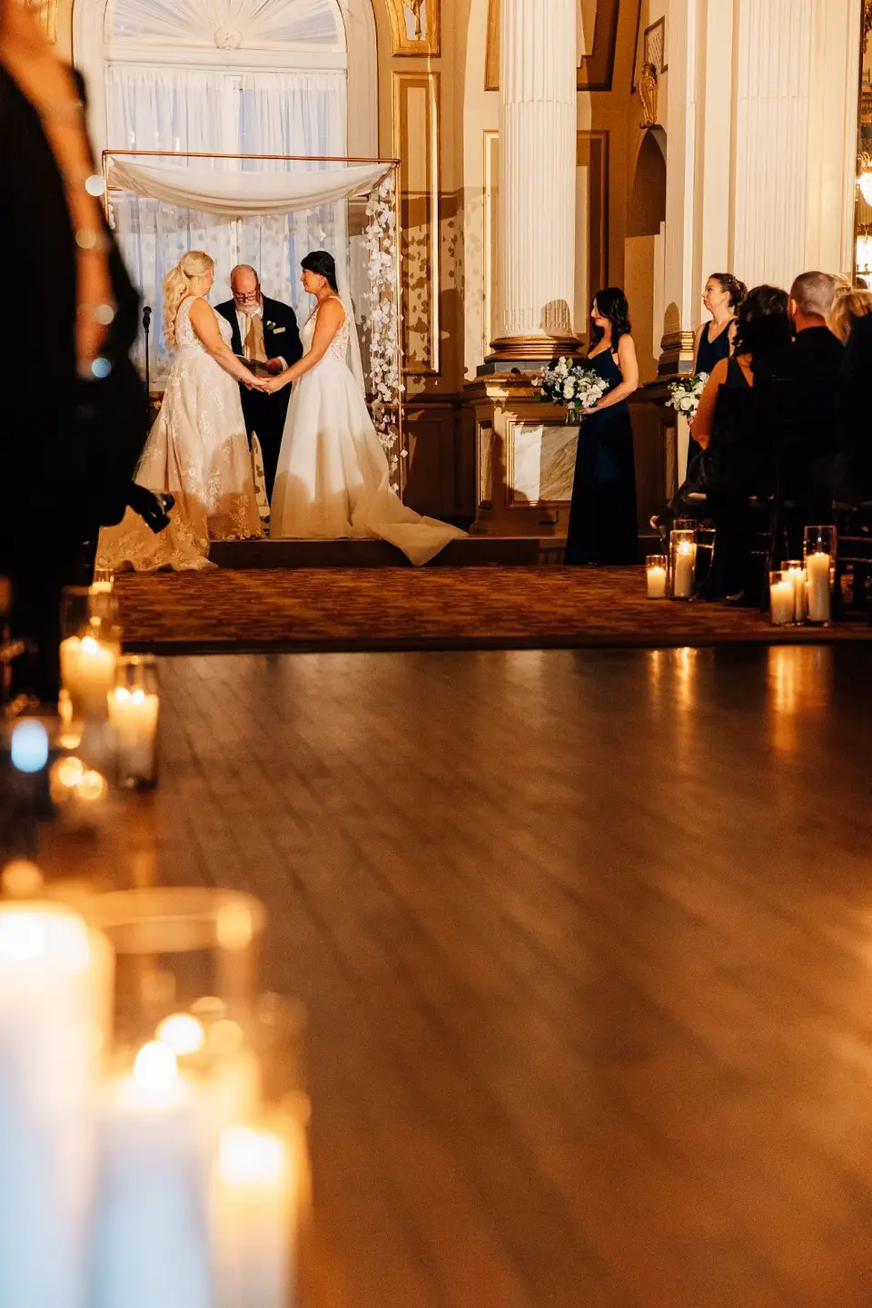 Two brides holding hands at the altar of a candlelit ballroom ceremony, seen from the aisle