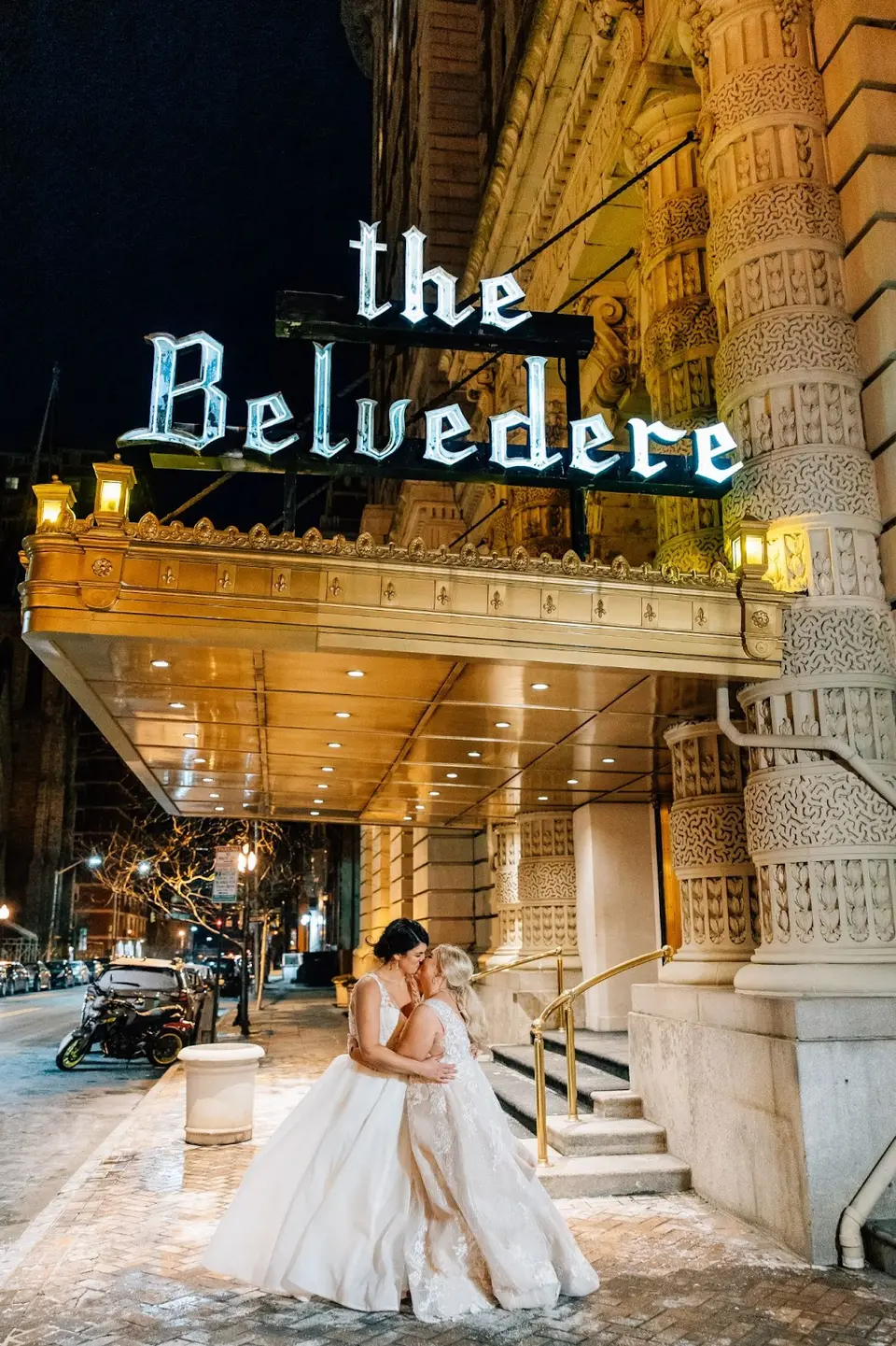 Two brides embracing under the lit Belvedere hotel sign at night