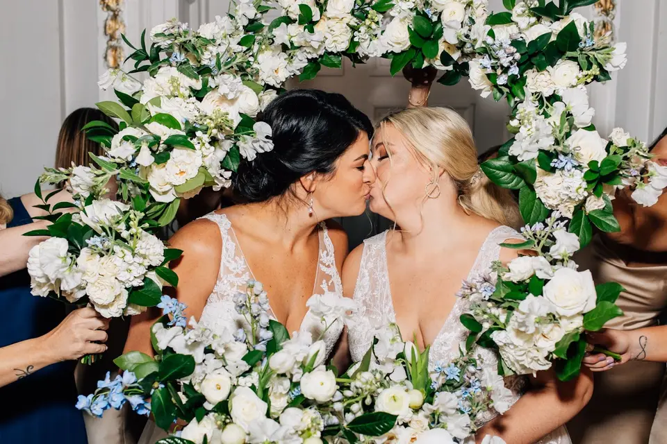 Two brides kissing while surrounded by white and blue floral bouquets
