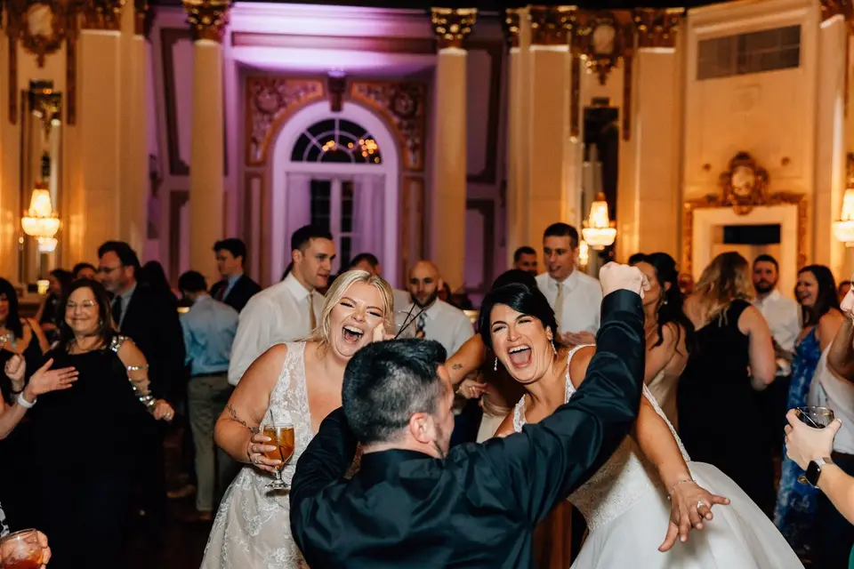 Two brides cheering and dancing with a guest on a crowded ballroom dance floor