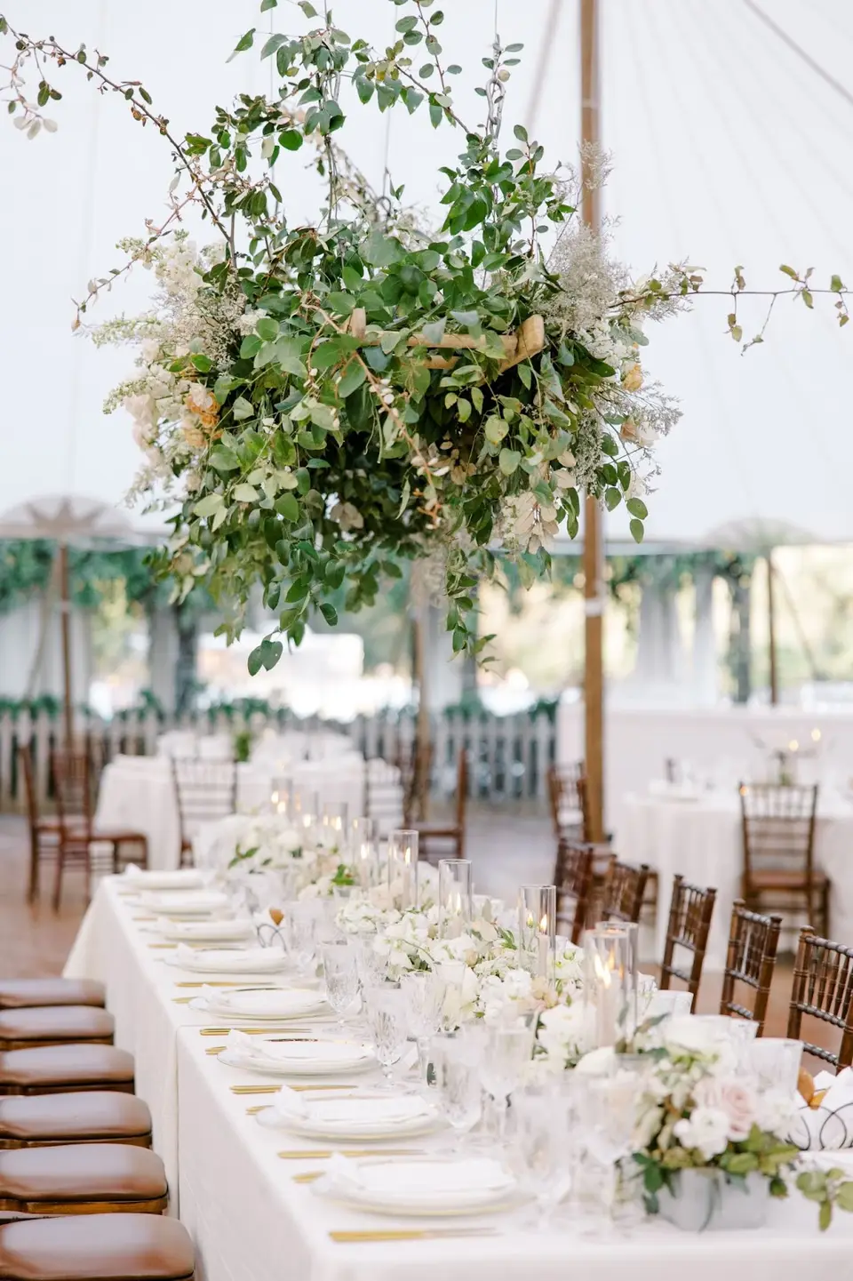 Long reception table with white linens, candles and flowers under a hanging greenery arrangement in a tent