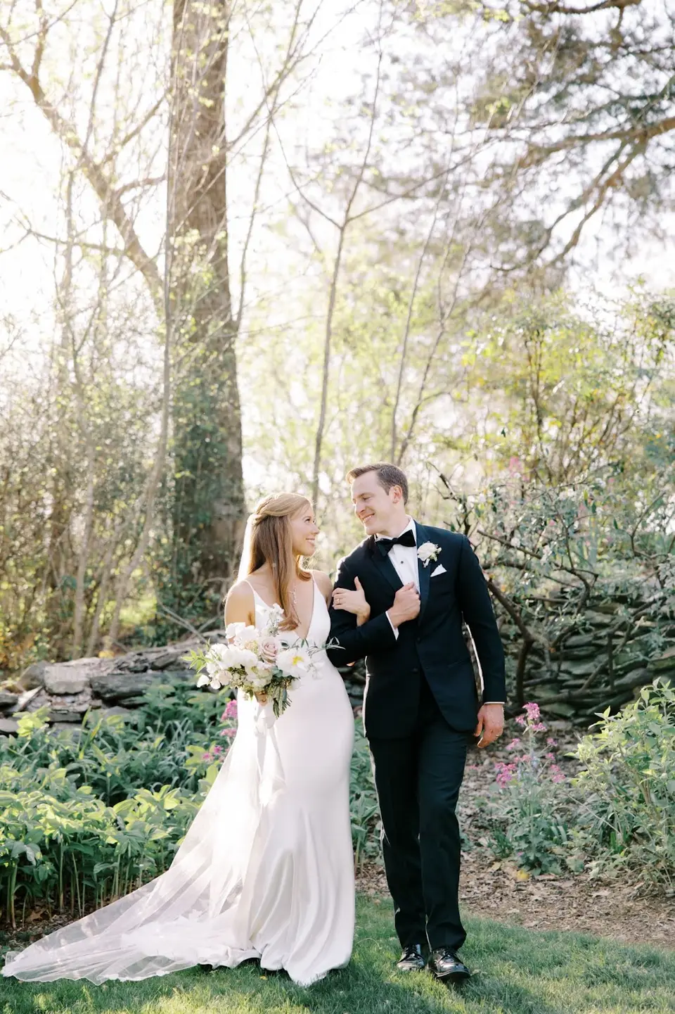 Bride in a white gown and groom in a black tuxedo walking arm in arm in a spring garden