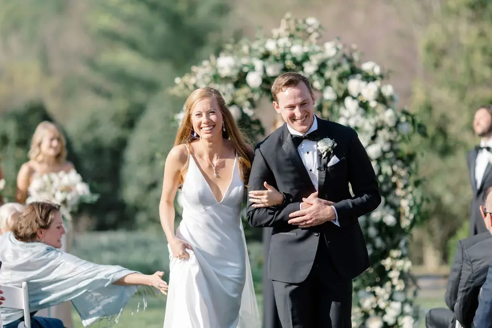 Smiling bride and groom walking up the aisle past a floral arch while a guest reaches toward them