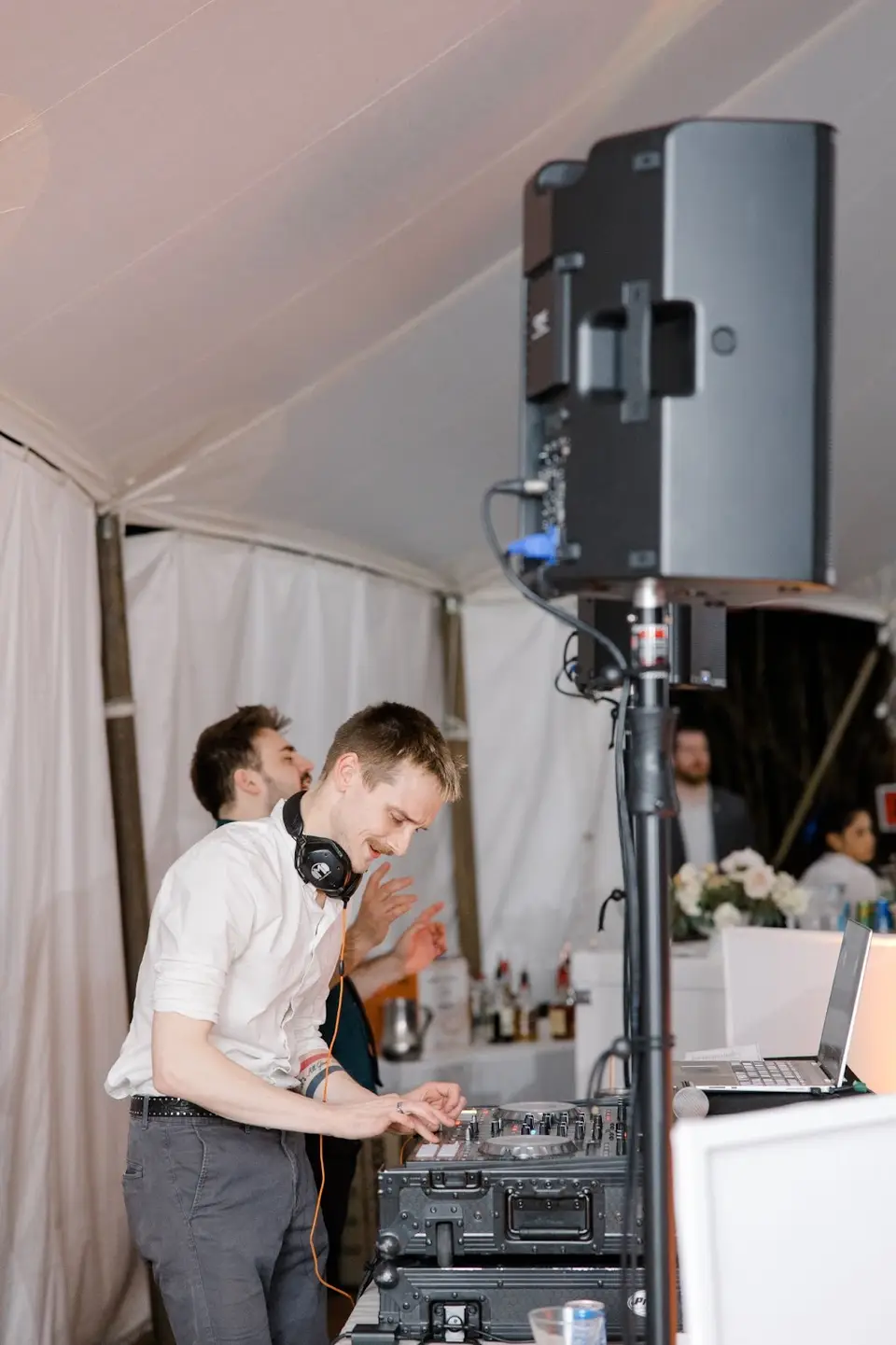 DJ working at a mixer and laptop beside a large speaker on a stand inside a white tent