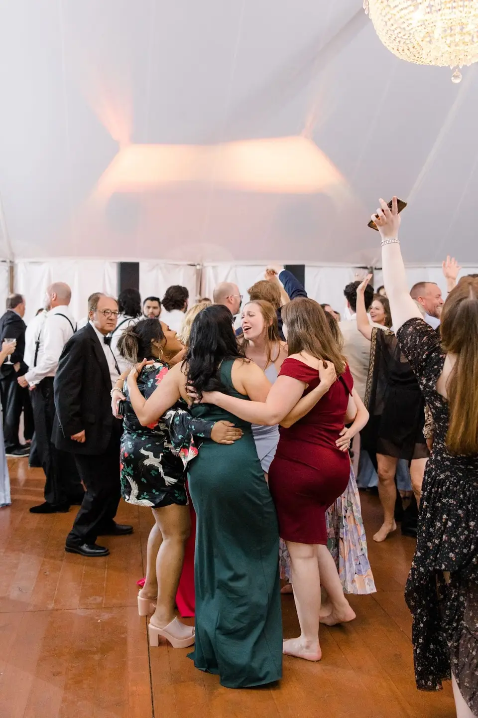 Women in colorful dresses hugging and dancing together on a wood dance floor under a white tent