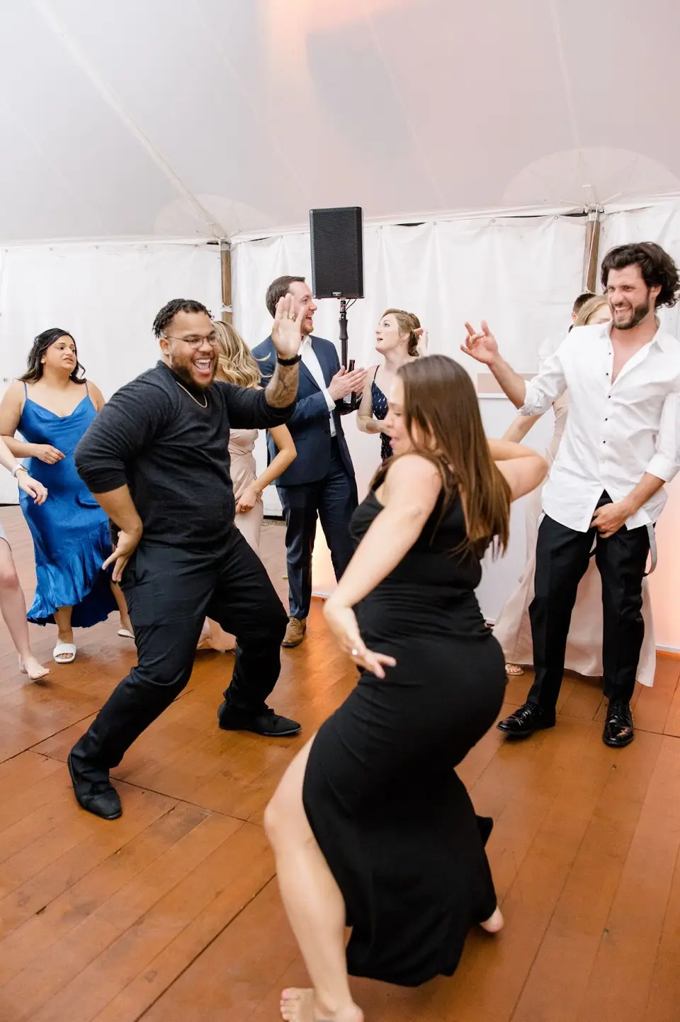 Guests dancing energetically on a wood floor under a white tent, with a man in black laughing
